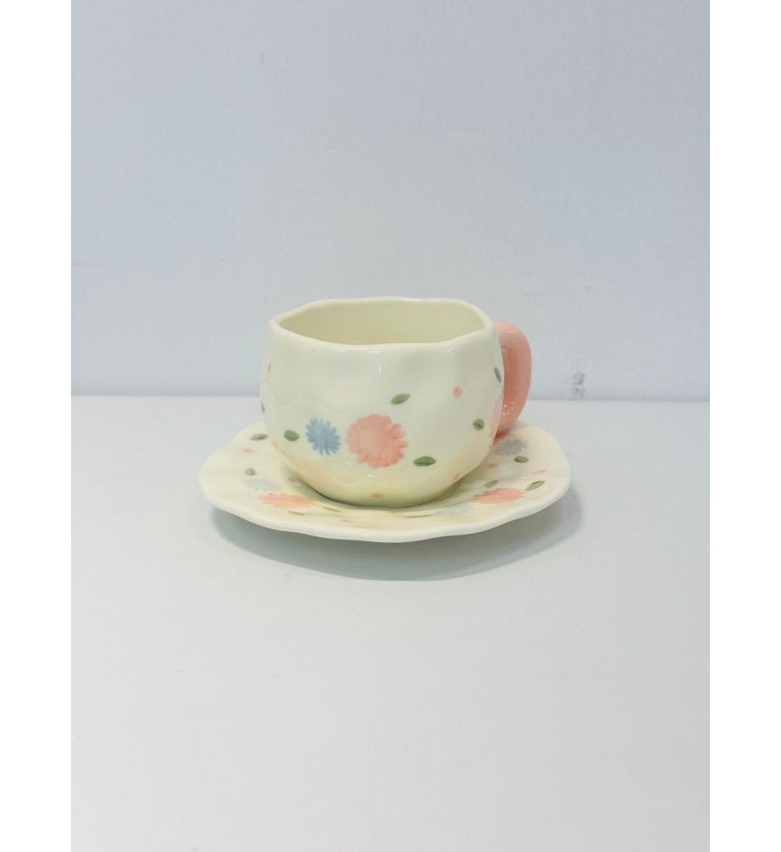 الكوب الظاهر في الصورة مصنوع من السيراميك بتصميم يدوي بنقوش زهرية بألوان هادئة مثل الوردي والخوخي والأزرق السماوي مع أوراق خضراء صغيرة