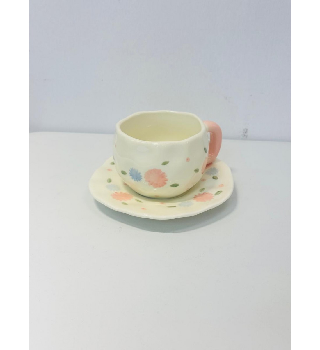 الكوب الظاهر في الصورة مصنوع من السيراميك بتصميم يدوي بنقوش زهرية بألوان هادئة مثل الوردي والخوخي والأزرق السماوي مع أوراق خضراء صغيرة